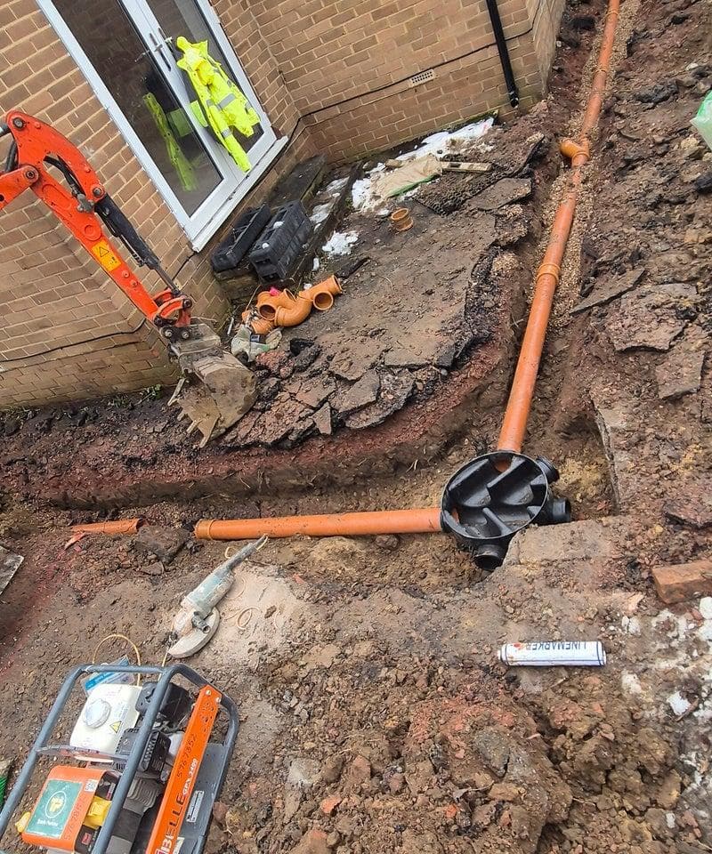Orange drainage pipes and a black inspection chamber in a trench near a brick house.